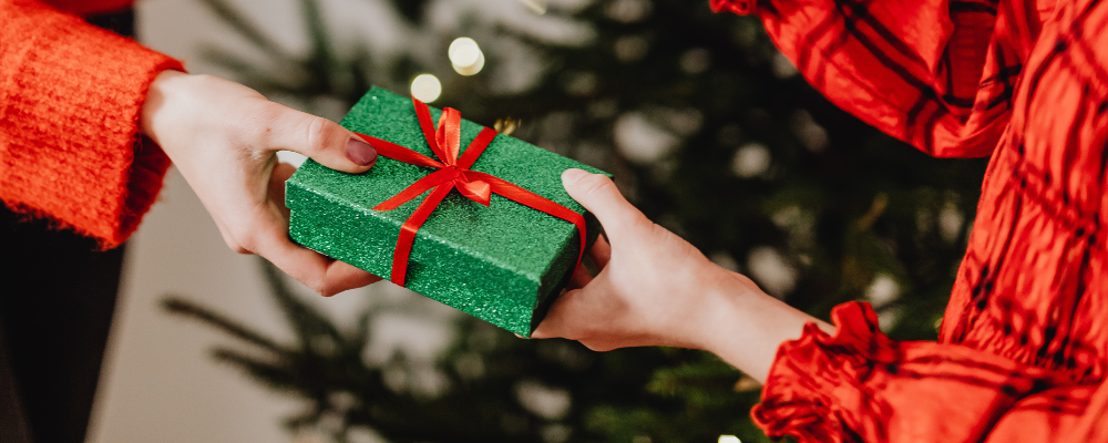 Wrapped green Christmas gift with a red ribbon being handed over in front of a Christmas tree