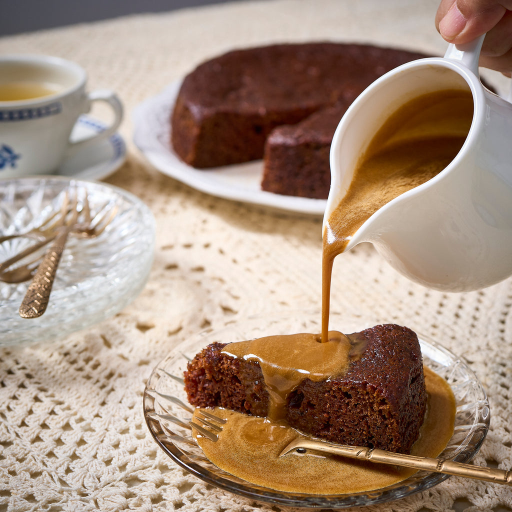Toffee sauce poured over a slice of Sticky Toffee Pudding, the whole pudding and a cup of tea behind
