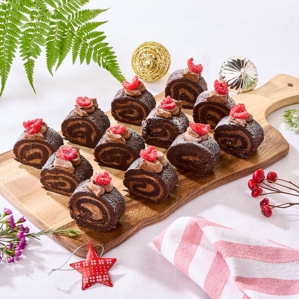 Twelve mini Christmas log cakes on a board, each topped with cream and a raspberry