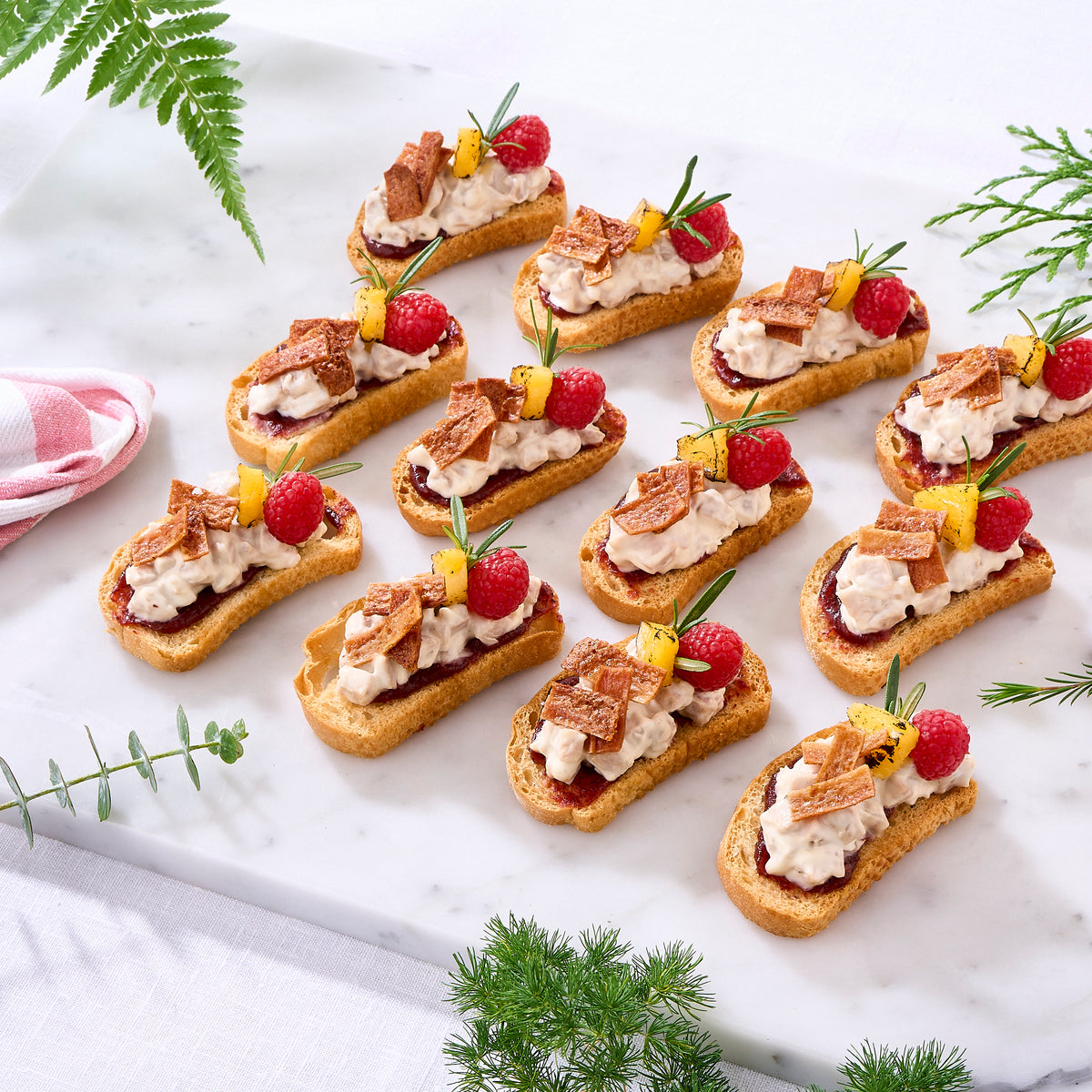 Six gourmet canapes on toasted bread, topped with white cream, dark berry preserves, chocolate curls, fresh raspberries, and pineapple slices, arranged on a white marble serving board with green leaves and a pink checkered cloth.