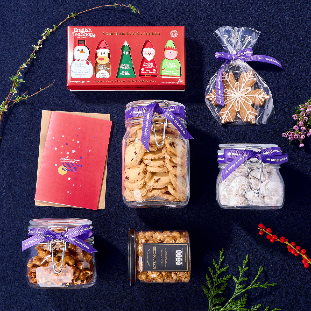 Flatlay of Christmas goodies including  glass jars of assorted cookies, set of organic teas, popcorn and a greeting card.